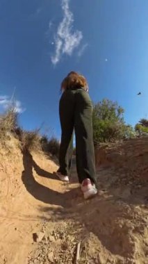 Dikey video. Toprak yolda yürüyen bir kadının dinamik hareketi ve yolu çevreleyen doğal bitkileri yakaladığı düşük açılı görüntüsü.