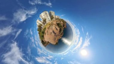 360 derecelik küçük bir gezegen efekti binalar, yeşillikler ve geniş mavi gökyüzü ile kayalık bir kıyı manzarasının dönüşünü yakalıyor ve gerçeküstü, küresel bir dünya perspektifi yaratıyor.