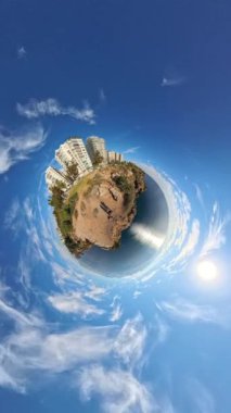 Dikey video. 360 derecelik küçük bir gezegen efekti binalar, yeşillikler ve geniş mavi gökyüzü ile kayalık bir kıyı manzarasının dönüşünü yakalıyor ve gerçeküstü, küresel bir dünya perspektifi yaratıyor.