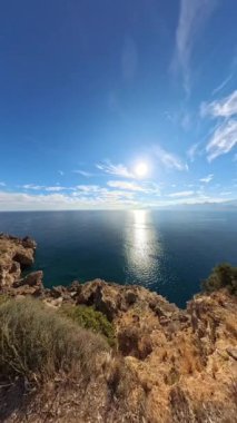 Dikey video. Güneşli bir okyanusun panoramik görüntüsü. Önünde kayalık bir uçurum ve uzaktan görülebilen dağlar. Parlak bir gökyüzü altında deniz manzarasının enginliğini yakalıyor..