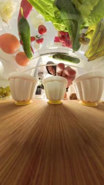 Buzdolabının içinden bir adamın kapıyı açtığını ve yeşil ile kırmızı jöle bardakları arasında gezindiğini görün. Geniş açılı çekim ön plandaki ahşap bir raftaki çeşitli yiyecekleri yakalar..