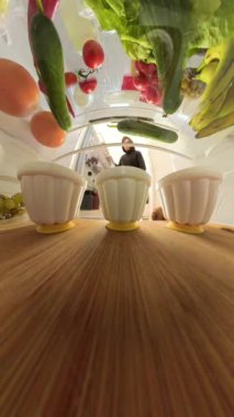 Buzdolabının içinde bir adam uzanır ve yeşil bir jöle kabı alır. Geniş açı perspektifi, yiyeceklerin ahşap bir rafta dizilişini yakalar ve seçim anını ön plana çıkarır..