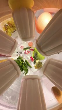 Geniş açı, yakınlaştırıcı etkisi olan bir buzdolabının içinde dönen manzara, konteynırlar, sebzeler ve meyveler gibi çeşitli ürünlerden giderek daha da uzaklaşır. Çekim, organize içeriği dinamik bir şekilde yakalar..