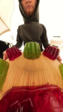 Buzdolabının içine sallanan kırmızı ve yeşil jöle bardakları yerleştiren bir adamın yakın çekim görüntüsü. Perspektif, ahşap bir raftaki jölenin canlı renklerini ve dokusunu vurgular..