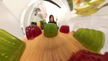 Buzdolabının içinden, bir adam sallanan kırmızı ve yeşil bir jöle tepsisi yerleştirir, yumuşak desenli jöleler rafta hafifçe sallanırken kapıyı kapatır..