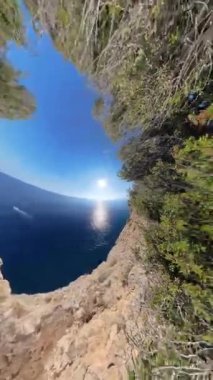 Dikey video. Okyanusla çevrili kayalık bir uçurumun küçük gezegen etkisi. Dönen bir kamera, çarpık bir perspektif yaratıyor. Güneş suyun üzerinde ışıl ışıl parlıyor, gerçeküstü, büyüleyici bir kalite ekliyor..
