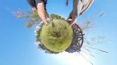 Küçük gezegen etkisi, çimenli bir parkta yürüyen bir çiftin bacaklarını gösteriyor, toprağı dairesel, gerçeküstü bir şekle dönüştürüyor, mavi gökyüzüne karşı..