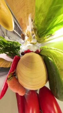 Dikey video. Bozuk bir perspektif, buzdolabının içinde domates, biber, marul ve havuç gibi taze sebzelerin ve sağlıklı ürünlerin raflarda canlı bir şekilde sıralandığını vurgular..