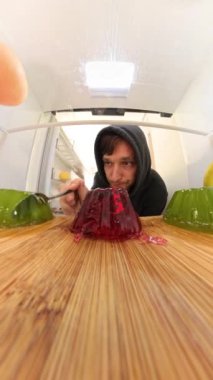 Dikey video. Buzdolabının içinden geniş açılı, zoom-out görüntüsü bir adamı kaşıkla kırmızı ve yeşil jöle yerken yakalar. Muz ve greyfurt gibi canlı meyveler sahneyi çerçeveler..