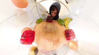 Balık gözü perspektifi, buzdolabının içinde muz ve greyfurt gibi meyvelerle kırmızı ve yeşil jöle yiyen bir adamın ahşap tepsiyi sardığını gösteriyor..