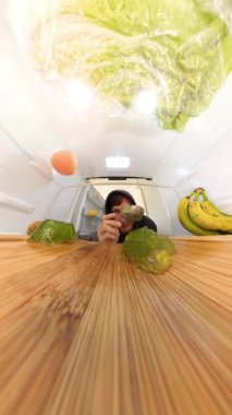 Dikey video. Dolly zoom buzdolabının içinde yeşil jöle yiyen bir adamı yakalar, ışık çok uzun süre sallanırken. Sahneyi kırmızı ve yeşil jöle, muz ve greyfurt çerçevesi.