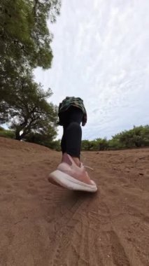 Dikey video. Bir kadın ağaçlarla çevrili kumlu bir patikada koşuyor, alçak açıyla çekiliyor. Yavaş çekim etkisi onun adımlarını ve bulutlu bir gökyüzünün altındaki doğal çevreyi vurguluyor..