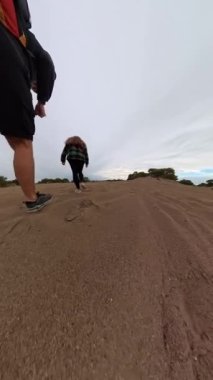 Dikey video. Ağır çekimde bir adam ve bir kadın çöl benzeri bir bölgede kumlu bir patikada yokuş yukarı yürüyorlar. Düşük açı görünümü onların adımlarını ve seyahat ederken kumun dokusunu vurgular..