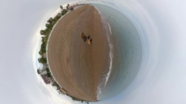 Bir Tiny Planet etkisi, bulutlu gökyüzü ve serin hava altında kumlu bir plajda yürüyen iki yolcuyu serin küresel bir manzara yaratarak serinletiyor..