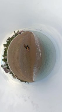Dikey video. Bir Tiny Planet etkisi, bulutlu gökyüzü ve serin hava altında kumlu bir plajda yürüyen iki yolcuyu serin küresel bir manzara yaratarak serinletiyor..