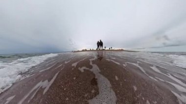 Dönen kamerayla çekilen yavaş çekim kumlu bir sahilde duran bir adamla bir kadını yakalar. Dalgalar kıyıya yavaşça yaklaşıp, kıyı şeridi ve gökyüzünün geniş açılı görüntüsüyle dinamik ve dinamik bir manzara yaratıyor..
