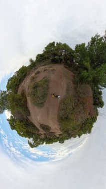 Dikey video. Küçük Gezegen etkisi, yeşil ağaçlar ve açık mavi gökyüzü ile çevrili bir çiftin yürüdüğü küresel bir orman manzarasını gösteriyor..