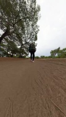 Dikey video. Ekose gömlekli bir kadın kumlu orman yolunda yürüyor, alçak bir açıdan yakalanmış, yumuşak zeminde ayak izleri var..