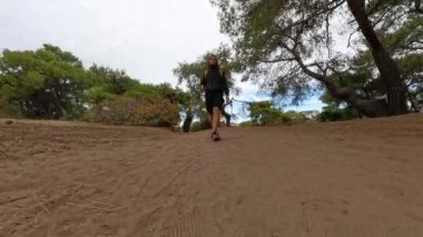 Kapüşonlu bir adam kumlu orman patikasında yürüyor tepesinde ağaçlar ve mavi gökyüzü var..