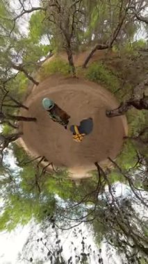 Dikey video. Çevresi yeşilliklerle çevrili eğri bir orman patikasında yürüyen bir çiftin küresel perspektifi yolu ve ağaçları aydınlatan küçük bir gezegen etkisi yaratıyor..