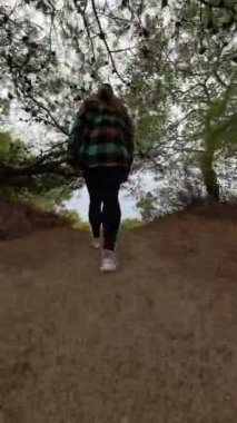 Dikey video. Bir kadın yüksek bir tepenin üstünde duruyor, ağaçların arasından uzak denize bakıyor. Dolly yakınlaştırma efekti onun bakış açısını ve sakin kıyı manzarasını vurgular.