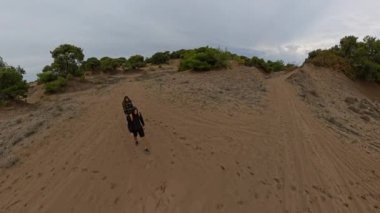 Etrafı çalılarla çevrili kumlu bir arazide yürüyen iki gezgin. Geniş açılı çekimler çorak araziyi vurguluyor..