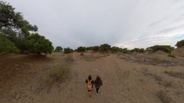 Uçsuz bucaksız bulutlu gökyüzünün altında dağınık yeşillikle kumlu bir alanda yürüyen iki yolcu. Arazinin açıklığını vurgulayan geniş açılı bir çekim..