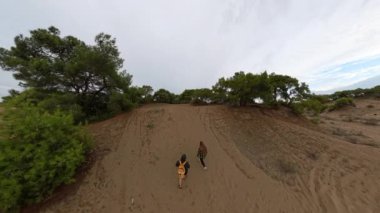 Erkek ve kadın kumlu bir yokuştan yükseliyor, yeşil çalılarla çerçevelenmiş ve yukarıdaki geniş bulutlu gökyüzü.