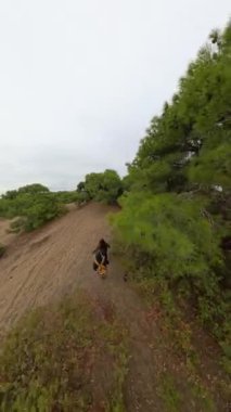 Dikey video. Bir adam ve bir kadın bulutlu bir gökyüzünün altındaki seyrek yeşil ağaçlarla çevrili kumlu arazide seyahat ediyorlar. Bakış açısı yukarıdan, açık ve ıssız ortamı gösteriyor..