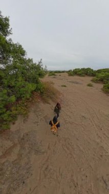 Dikey video. Kamera sarı çantalı adama odaklanıyor ve kumlu bir patikada yürüyen kadına. Etrafı seyrek yeşilliklerle çevrili, yukarıdan bakan kadına..