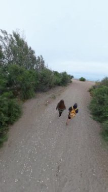 Dikey video. Etrafı çalılarla çevrili kumlu bir patika boyunca yürüyen iki kişi bulutlu bir gökyüzünün altında sahile doğru ilerliyorlar. Bakış açısı geniş ve hafifçe yükseltilmiş..