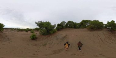 Havadan bakış açısı. Hava perspektifinden bakıldığında 360 derecelik pürüzsüz bir VR panoraması, sakin bir açık hava macerasında bulutlu bir gökyüzüne karşı uzanan kumlu bir arazide çalılar ve küçük ağaçlarla yürüyen iki gezgini yakalar..