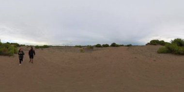 Kusursuz bir 360 derecelik küresel VR panoraması, bulutlu bir gökyüzünün altında seyrek bitki örtüsüyle kumlu bir arazide yürüyen iki gezgini yakalar ve barışçıl ve sürükleyici bir perspektif sunar..