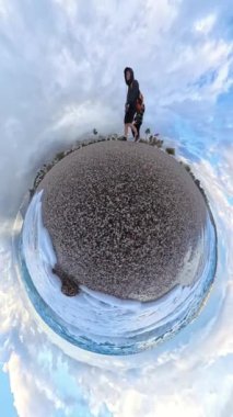 Dikey video. Ağır çekim. Okyanusla çevrili bir çakıl taşlı plajda gezinen bir çiftin küçük gezegen görüntüsü, dramatik bir bulut karışımı ve parlak mavi gökyüzü gerçeküstü bir atmosfer yaratıyor..