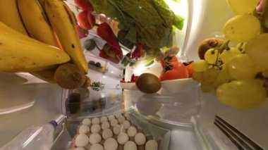 Buzdolabının içinden bir perspektif, canlı ve doğal bir ortamda renkli meyve ve sebzelerle çevrili bir adamın kapıyı açtığını ve bir çilek yediğini gösteriyor..