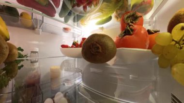 Buzdolabının içinden eşsiz bir perspektif, iyi aydınlatılmış bir mutfak ortamında renkli meyve ve sebzelerle çevrili bir adamın kiviye uzandığını gösteriyor..