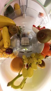 Dikey video. Buzdolabının içinden küresel bir manzara, canlı meyve ve sebze çeşitleriyle çevrili bir kivi alan bir adamın sanatsal bir mutfak sahnesi yarattığını gösteriyor..