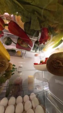 Dikey video. Buzdolabının içinde dönen bir kamera kapıyı açan ve canlı meyve ve sebzelerle çevrili bir kiviye uzanan bir adamı yakalar ve dinamik ve sürükleyici bir sahne yaratır..