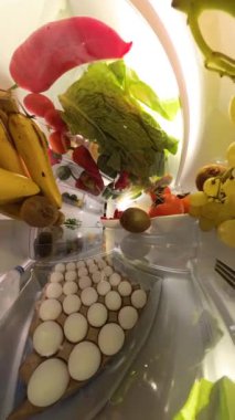 Dikey video. Buzdolabının içinden bir perspektif, canlı ve doğal bir ortamda renkli meyve ve sebzelerle çevrili bir adamın kapıyı açtığını ve bir çilek yediğini gösteriyor..