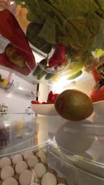 Dikey video. Buzdolabının içinden yakından bakınca, canlı meyve ve sebzelerle çevrili, parlak ve taze bir tabaktan çileğe uzanan bir adam görülüyor..