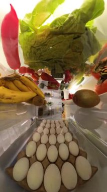 Dikey video. Doli zum efekti buzdolabını açan ve canlı meyve ve sebzelerle çevrili çilek yiyen bir adamı parlak ve taze bir iç mekanda yakalar..