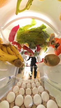 Dikey video. Doli zum efekti, rengarenk meyveler ve canlı ürünlerle çevrili buzdolabından salata için çeşitli sebzeler alan bir adamı parlak ve taze bir şekilde yakalar..