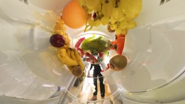 Dolly zoom, taze meyve ve renkli ürünlerle çevrili buzdolabından salata için çeşitli sebzeler alan bir adamı canlı ve büyüleyici bir sahnede yakalar..