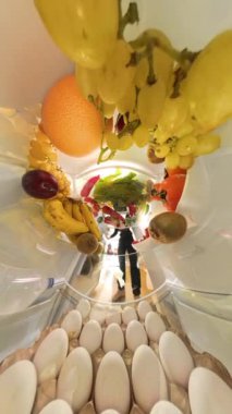 Dikey video. Dolly zoom, taze meyve ve renkli ürünlerle çevrili buzdolabından salata için çeşitli sebzeler alan bir adamı canlı ve büyüleyici bir sahnede yakalar..