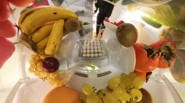 Buzdolabının içindeki küresel bir manzara, muz, kivi ve üzümler de dahil olmak üzere taze ürünlerle çevrili bir kutudan yumurta toplayan bir adamı dinamik ve daldırıcı bir sahne yaratırken yakalar..