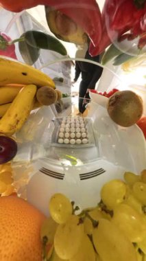 Dikey video. Buzdolabının içindeki küresel bir manzara, muz, kivi ve üzümler de dahil olmak üzere taze ürünlerle çevrili bir kutudan yumurta toplayan bir adamı dinamik ve daldırıcı bir sahne yaratırken yakalar..