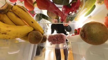 Buzdolabının içinden bir perspektif, canlı meyve ve sebzelerle çevrili et konteynırına uzanan bir adamın sebzeden ete geçişi vurguluyor..