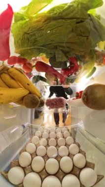 Dikey video. Buzdolabının içinden bir perspektif, canlı meyve ve sebzelerle çevrili et konteynırına uzanan bir adamın sebzeden ete geçişi vurguluyor..