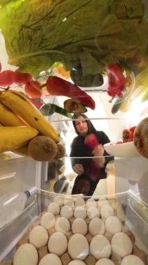 Dikey video. Yakın plan bakış açısı, taze ürünlerle çevrili buzdolabını açtıktan sonra et konteynırını inceleyen bir adamın sebzeler yerine eti seçme kararını sembolize ettiğini gösteriyor..