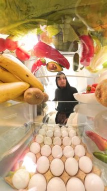 Dikey video. Buzdolabından alınmış renkli meyve ve sebzelerle çevrili bir et paketini inceleyen bir adam, vejetaryen seçeneklerine karşı görsel olarak bir seçim sunuyor..